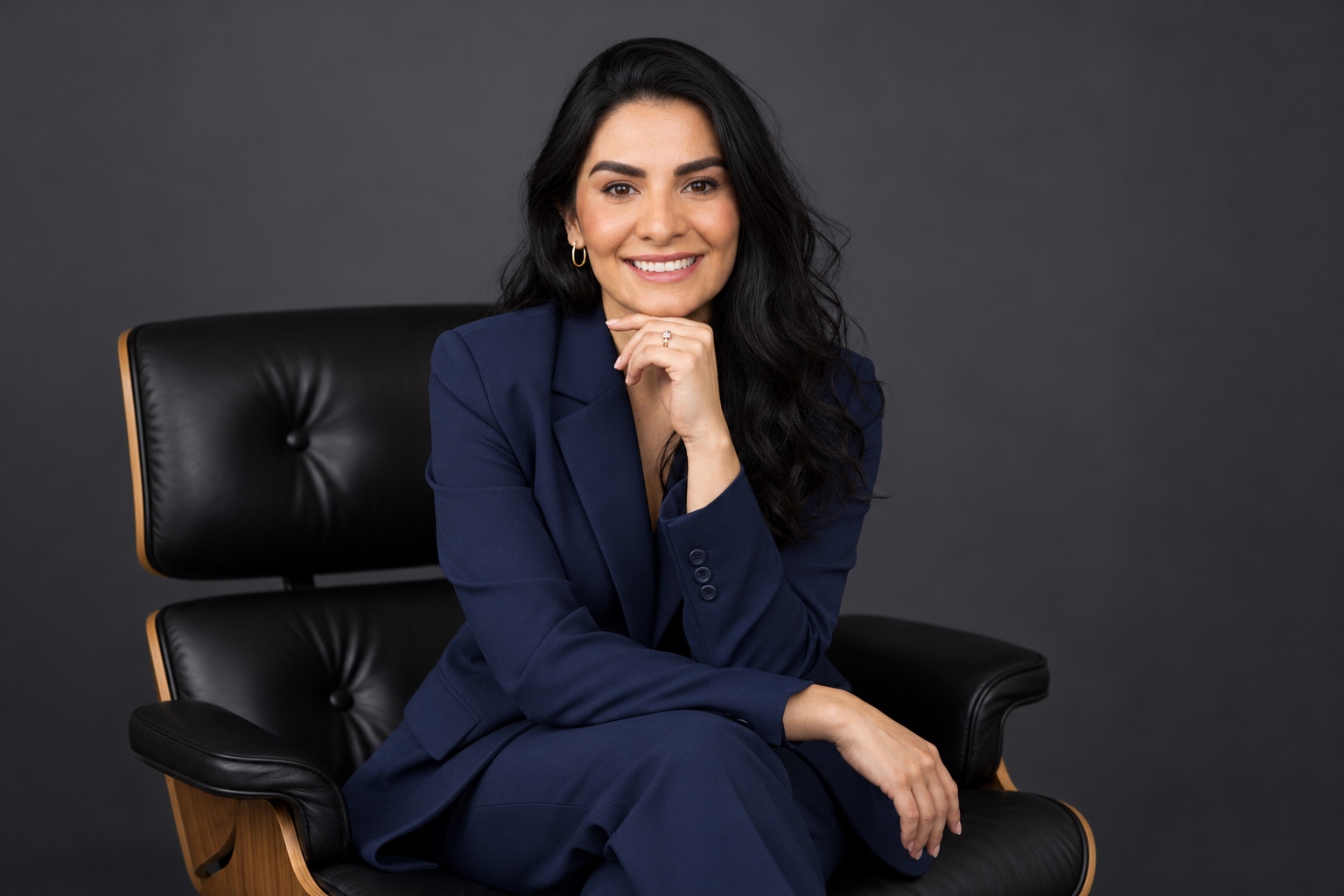 Daniele Noni, de terno azul-marinho, sentada em uma poltrona de couro, sorrindo com a mão apoiada no queixo.